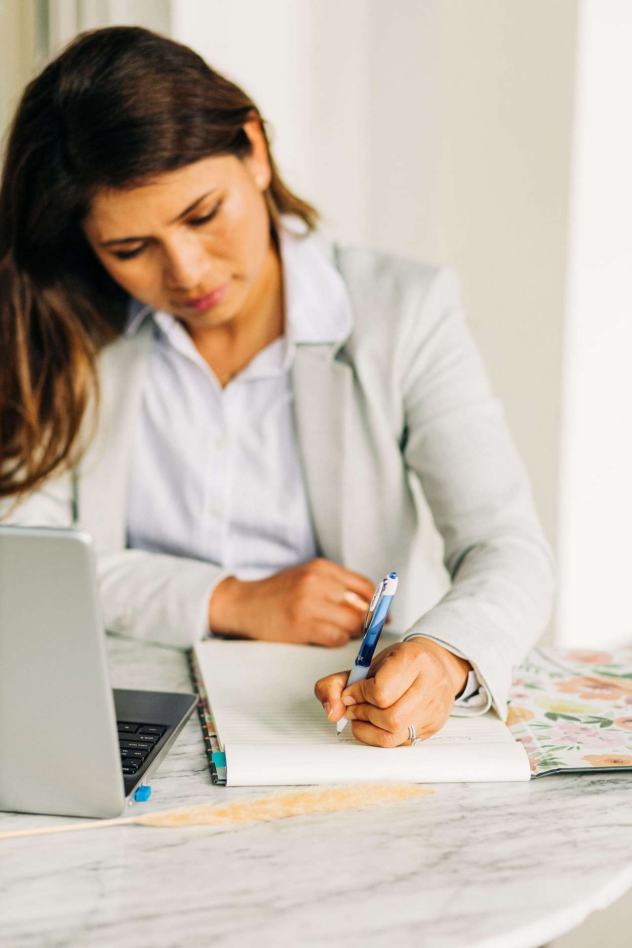 A person in a light gray blazer is seated at a desk, writing with clarity in a notebook using a blue pen. An open laptop, strategically positioned to their left, highlights their sense of direction.